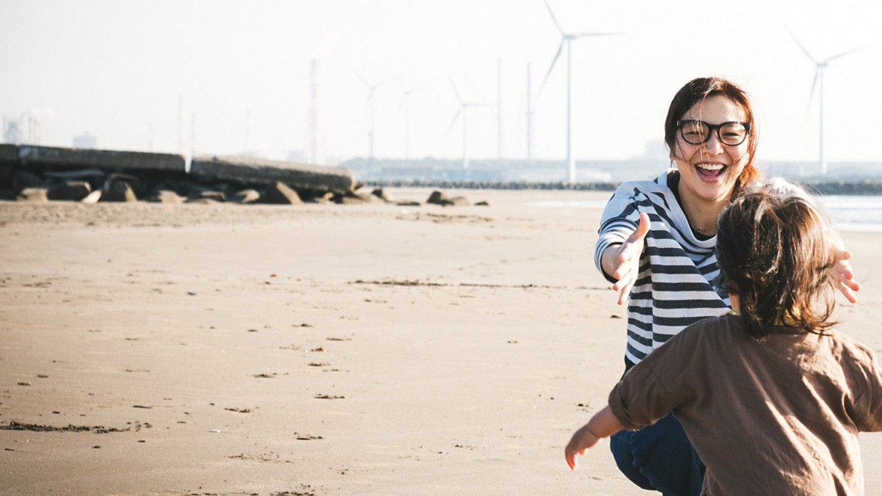 Vaikas paplūdimiu bėga pas mamą, fone – vėjo jėgainės